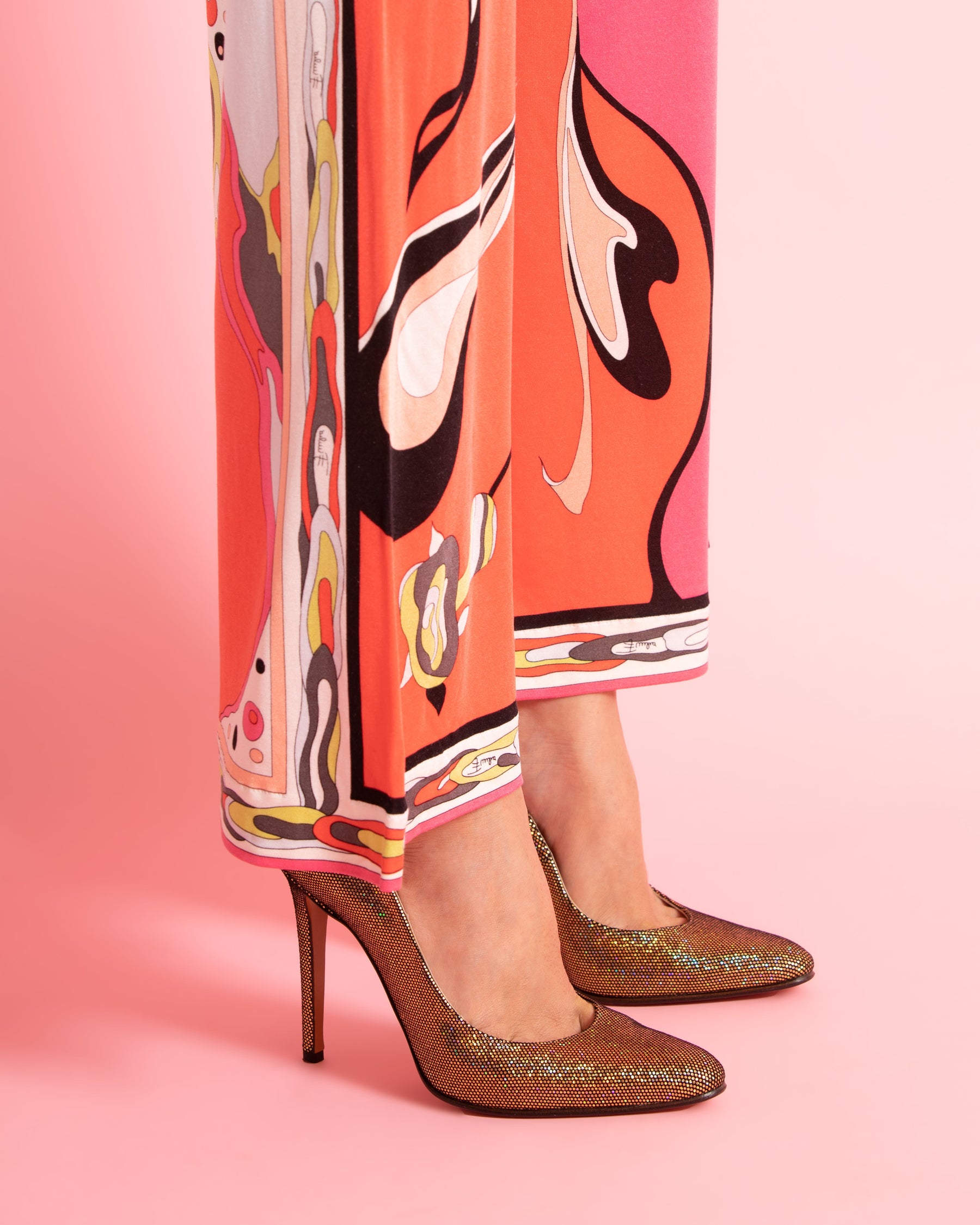 Close-up of a person wearing multicolored, abstract-patterned pants and sparkling gold high heels on a pink background, conveying a bold, stylish tone.